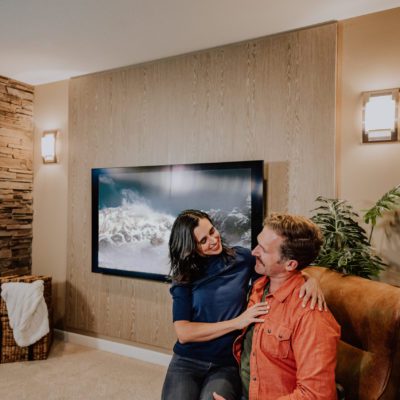 A couple in their media room with an acoustic wood wall, stone fireplace, television, and a leather chair.