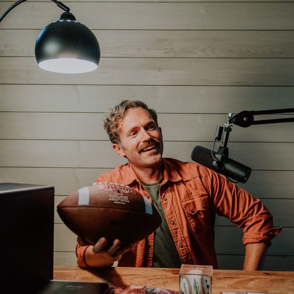 Man holds football in his podcast room in front of maple wood wall.